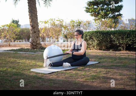 Femme enceinte en vêtements de sport noirs se trouve sur un tapis de yoga à l'extérieur, en utilisant une bande de résistance pour un entraînement en force douce. Une boule de stabilité repose à proximité Banque D'Images