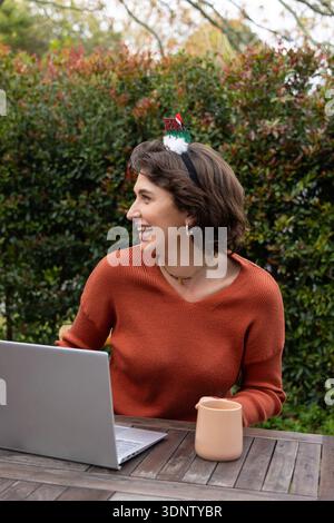 femme des années 20 portant un pull de rouille et un bandeau festif, riant à la table d'arrière-cour avec tasse d'ordinateur portable Banque D'Images