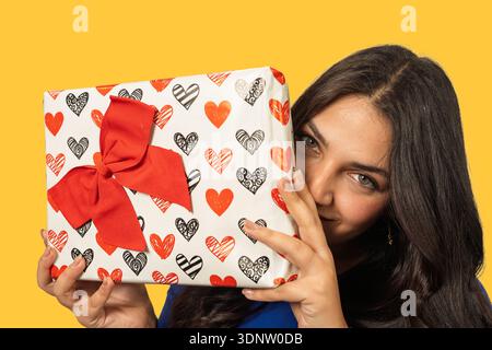 Femme jetant un coup d'œil de derrière une boîte cadeau avec des motifs de coeur rouge pour une occasion spéciale Banque D'Images