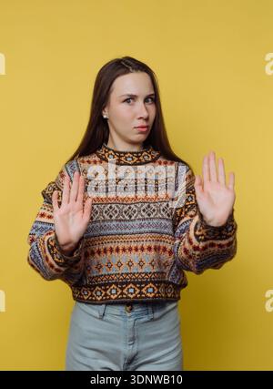Une jeune femme se tient confiante avec les paumes levées, signalant de s'arrêter. Son pull aux motifs éclatants contraste magnifiquement avec le joyeux YE Banque D'Images