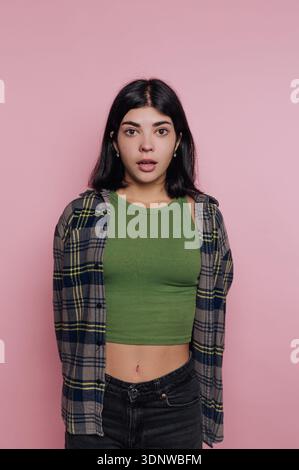Une jeune femme se tient contre un mur rose doux, portant un short top vert et une chemise à carreaux ouverte. Son expression surprise capture un moment de spontan Banque D'Images
