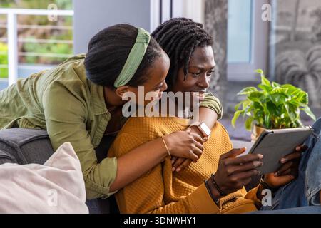 Couple afro-américain assis sur un canapé gris, homme tenant une tablette, femme penchée et embrassant Banque D'Images