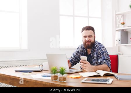 Jeune homme d'affaires souriant dans le bureau en utilisant le téléphone portable par l'ordinateur portable Banque D'Images