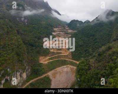 Une vue aérienne montre une route de montagne sinueuse coupant à travers des pentes abruptes et boisées avec des roches exposées et des travaux de terrassement, partiellement enveloppée dans un brouillard bas. Banque D'Images