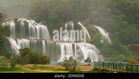 La cascade de Ban Gioc coule au-dessus de larges cascades étagées dans une rivière calme, avec une passerelle piétonne et des ruisseaux peu profonds au premier plan. Banque D'Images