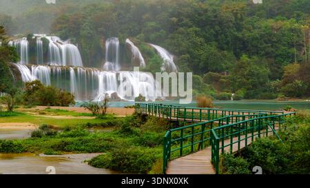 La cascade de Ban Gioc se déverse en larges niveaux dans une rivière turquoise, avec une passerelle piétonne courbe traversant des ruisseaux peu profonds. Banque D'Images
