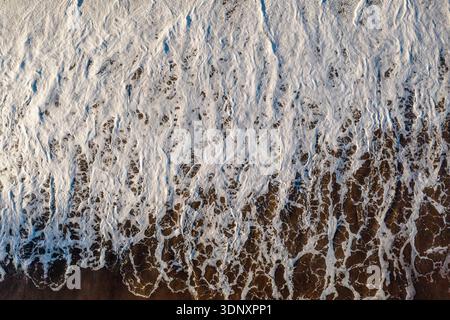 Vue de dessus des vagues mousseuses caressant doucement le rivage sablonneux d'une plage minière dans les Asturies, Espagne. Les motifs uniques créent une saisonnière abstraite fascinante Banque D'Images