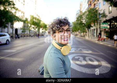 Une femme adulte joyeuse avec des cheveux bouclés et des lunettes apprécie une promenade en ville. Portant des écouteurs, elle adopte le style et la technologie modernes tout en exprimant p Banque D'Images
