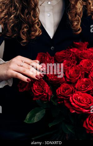 Une femme aux cheveux bouclés tient doucement un beau bouquet de roses rouges vibrantes. Ses ongles manucurés complètent les pétales rouges profonds, créant une frappe Banque D'Images