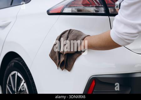 Un jeune homme nettoie soigneusement et entretient sa voiture électrique blanche, assurant une apparence impeccable et impeccable. Cette image dépeint la vie moderne Banque D'Images
