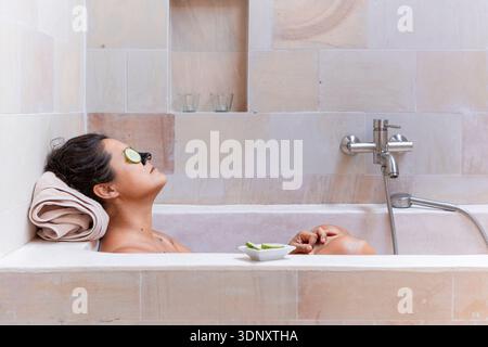 Une femme se détend dans une baignoire contemporaine, profitant d'un moment de sérénité avec des tranches de concombre sur ses yeux. Le cadre reflète un accent mis sur les soins personnels et Banque D'Images