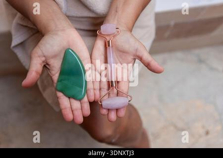 Gros plan des mains tenant un rouleau facial en quartz rose et un outil de jade. Ces pierres de beauté faites à la main sont utilisées dans les routines de soins de la peau pour favoriser la relaxatio Banque D'Images