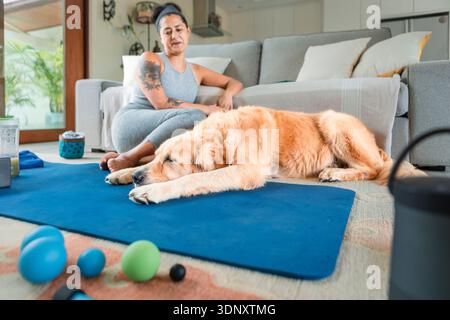 Une femme en tenue athlétique est assise sur un tapis de yoga à l'intérieur, se relaxant après l'exercice. À côté d'elle, un Golden retriever repose confortablement. Divers accès au centre de fitness Banque D'Images