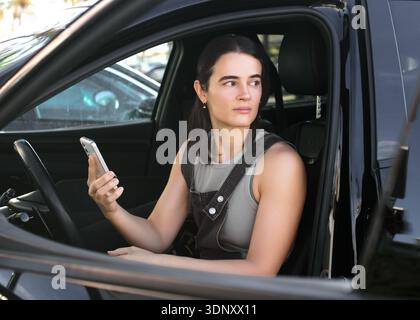 Une femme est assise dans sa voiture garée, regardant soigneusement par la fenêtre tout en tenant un smartphone. La lumière du soleil filtre à travers, mettant en évidence son expre serein Banque D'Images
