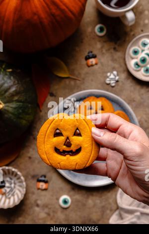 Vue de dessus d'une personne rognée méconnaissable tenant un biscuit d'Halloween en forme de citrouille avec du chocolat. La table rustique présente plus de biscuits, citrouilles Banque D'Images
