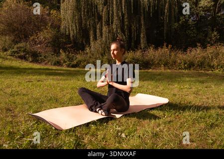 Une jeune femme pratique le yoga en plein air, assise dans une pose méditative sur un tapis. Elle est entourée de verdure naturelle, profitant d'un moment paisible de Banque D'Images