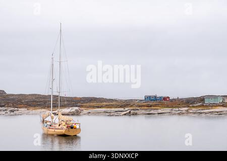 Une scène sereine au Groenland présente un voilier doucement ancré près du rivage rocheux, avec de petites cabines colorées peintes en bleu, rouge et vert Banque D'Images