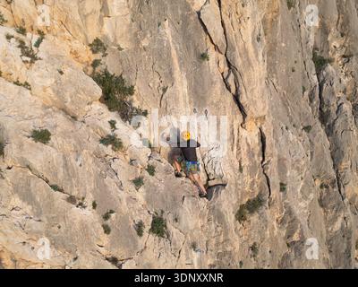 Grimpeur avec un casque et une corde escaladant une falaise accidentée sur la Costa Blanca. La formation rocheuse et les taches vertes soulignent l'aventure palpitante en plein air Banque D'Images