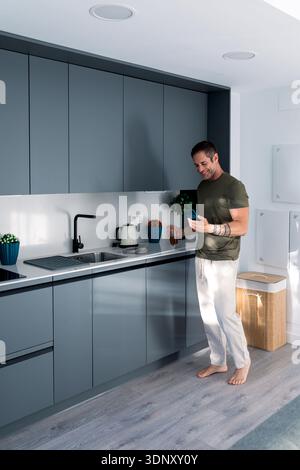 Un homme se tient pieds nus dans une cuisine moderne et élégante, tenant un smartphone et dégustant une boisson. La cuisine dispose d'armoires gris foncé et d'un lumineux, un Banque D'Images
