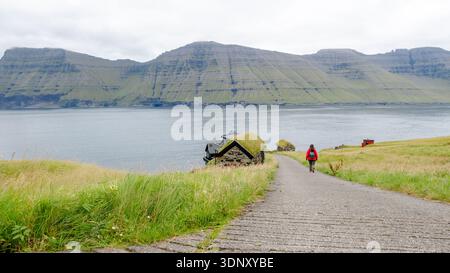 Une femme en veste rouge marche le long d'un chemin pavé à Mikladalur, Kalsoy, îles Féroé, avec des maisons à toit d'herbe et des montagnes escarpées en arrière-plan Banque D'Images