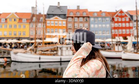 Une femme élégante dans un haut fleuri et un chapeau regarde les bâtiments colorés du XVIIe siècle et les bateaux qui bordent le canal Nyhavn à Copenhague, au Danemark, capturant t Banque D'Images