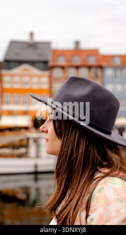 Une femme réfléchie portant un chapeau élégant regarde au loin tout en se tenant près des bâtiments pittoresques et colorés de Nyhavn, Copenhague, sur an Banque D'Images