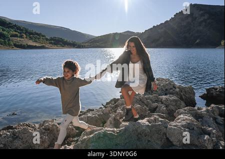 Un moment joyeux d'une mère et de sa fille se tenant la main et explorant le rivage rocheux d'un lac italien paisible par un après-midi ensoleillé, avec mountai Banque D'Images