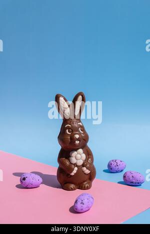 Un charmant lapin de Pâques en chocolat avec une fleur blanche, se dresse au milieu d'œufs mouchetés violets. Situé sur un fond rose pastel et bleu, la scène incarne Banque D'Images
