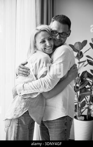 Couple heureux embrassant sur fond clair à la maison. Jolie femme souriant et embrassant son mari. Couple doux en vêtements décontractés. Image en noir et blanc. Banque D'Images