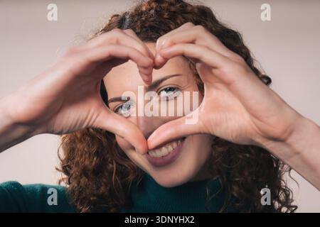 Une jeune femme aux cheveux roux bouclés sourit et forme une forme de cœur avec ses mains, exprimant amour et soin. Banque D'Images