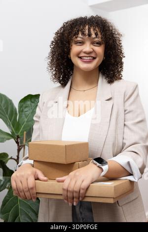 Femme afro-américaine souriante et tenant trois boîtes d'expédition au bureau portant un blazer beige Banque D'Images