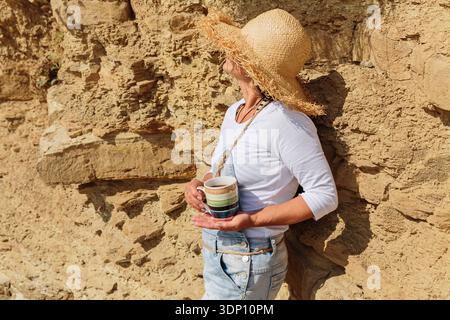Femme dans le chapeau de paille se relaxant avec du café par la côte rocheuse Banque D'Images