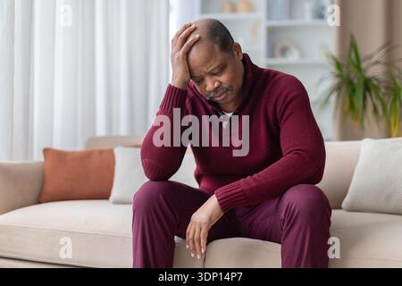 Homme noir mature assis sur le canapé avec la main sur la tête, regardant inquiet Banque D'Images