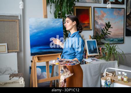 Artiste femme positive peignant sur toile à l'appartement confortable loft, plein d'idées et d'inspiration. Banque D'Images
