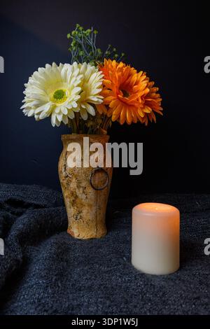 Nature morte discrète dramatique avec des fleurs de gerbera orange et blanches dans un vase rustique, bougie allumée sur un tissu sombre, ambiance de soirée confortable et espace de copie. Banque D'Images