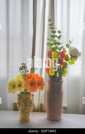 Deux vases avec des tulipes, des fleurs d'eucalyptus et de gerbera par des rideaux transparents, une nature morte intérieure douce rétro-éclairée avec une ambiance calme et un espace copie. Banque D'Images