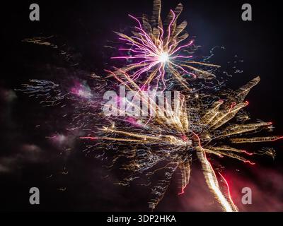 Explosion de feu d'artifice doré en bas à droite, éclatement coloré plus petit avec noyau blanc brillant au-dessus, lueur rouge s'estompant à droite, nuit noire profonde percée par la lumière. Banque D'Images