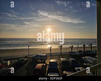 USAGE ÉDITORIAL EXCLUSIF - DAYTONA BEACH FLORIDA : une vue sereine sur le coucher du soleil sur l'océan Atlantique depuis un balcon en bord de mer en Floride. La photo capture le soleil qui se reflète sur les vagues de l’océan, une plage de sable bordée de palmiers et un parking côtier au premier plan pendant l’heure dorée. Banque D'Images