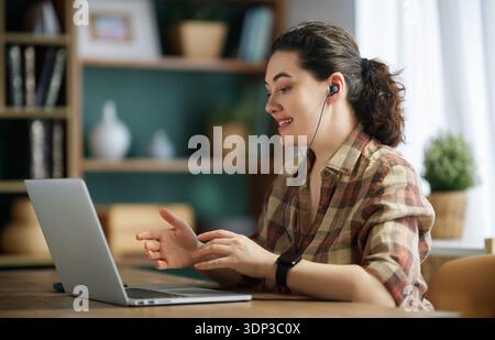 Une femme professionnelle participe à une vidéoconférence depuis son bureau à domicile confortable. Elle sourit et porte des écouteurs, incarnant une baguette à distance productive Banque D'Images