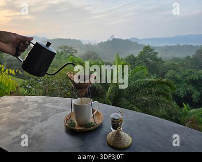 Vivez des moments sereins avec une tasse de café fraîchement moulu tout en surplombant la magnifique Khao Sok Thaïlande. Une végétation luxuriante et des montagnes brumeuses offrent le cadre idéal pour la détente. Banque D'Images