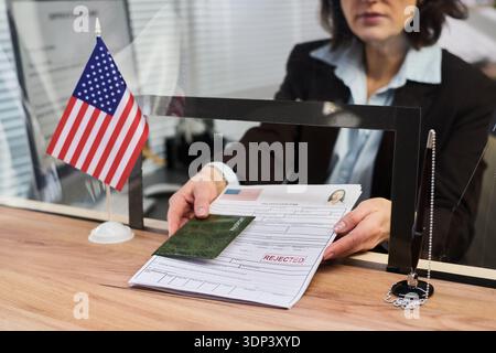 Femme caucasienne d'âge moyen traitant la demande de visa au comptoir, tenant des documents avec timbre et passeport rejetés, drapeau américain sur le bureau, travaillant dans le centre de visa Banque D'Images