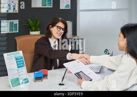 Femme caucasienne d'âge moyen assise au bureau serrant la main avec une jeune femme latine adulte, tout en examinant les documents de demande de visa dans le cadre moderne de bureau de centre de visa Banque D'Images