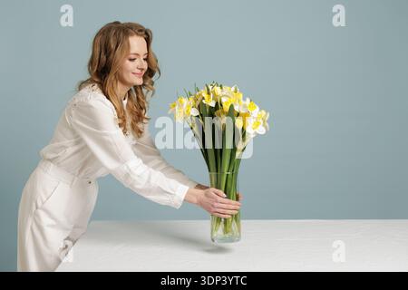Femme arrangeant des jonquilles dans un vase en verre sur la table. Idéal pour la promotion de la floraison, l'image de marque de style de vie et les campagnes de marketing printanières. Banque D'Images