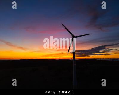 La vue aérienne capture une éolienne moderne au coucher du soleil avec une lumière chaude et un ciel spectaculaire mettant en valeur les infrastructures d'énergie renouvelable. Banque D'Images