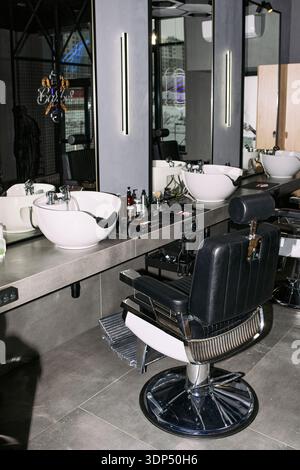 Station de coiffure vide avec chaise de barbier moderne positionnée devant un grand miroir avec évier de lavage de cheveux professionnel et divers outils de toilettage, disposés sur le comptoir Banque D'Images