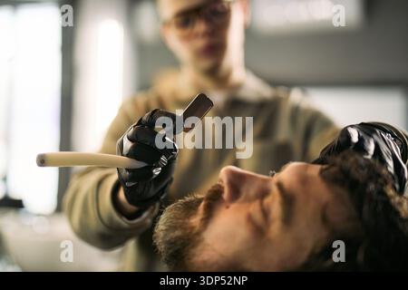 Jeune homme adulte caucasien incliné tandis que jeune homme adulte caucasien coiffeur portant des gants préparant un rasoir droit pour le rasage dans un salon de coiffure, concentrez-vous sur la main du barbier et le rasoir Banque D'Images
