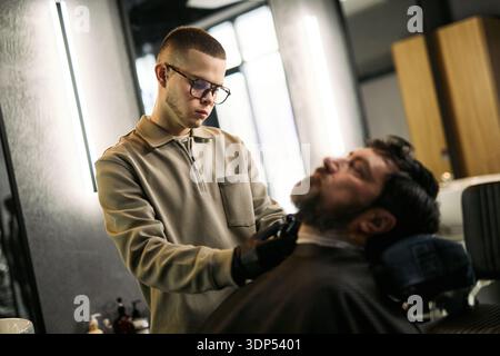 Jeune homme caucasien adulte portant des lunettes taillant la barbe de l'homme caucasien d'âge moyen couché dans la chaise de barbier, se concentrant sur le processus de toilettage dans le cadre moderne de barbier Banque D'Images