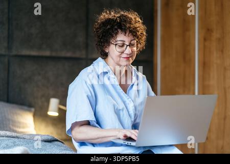 Femme blanche calme dans ses années 40 avec lunettes tapant sur ordinateur portable dans la chambre, lumière chaude de lampe Banque D'Images