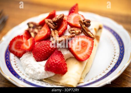 Une crêpe pliée sur une assiette blanche surmontée de fraises tranchées, de pacanes et de fromage cottage sur une surface de table en bois. Banque D'Images