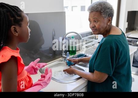 Grand-mère féminine senior et petit-enfant se lavant à l'évier de cuisine, enfant dans des gants roses avec éponge bleue Banque D'Images